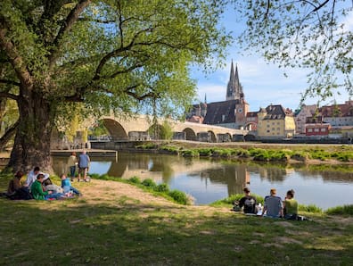 Donauufer in Regensburg, wo Leute im Gras sitzen, mit Blick auf die Altstadt und eine Brücke